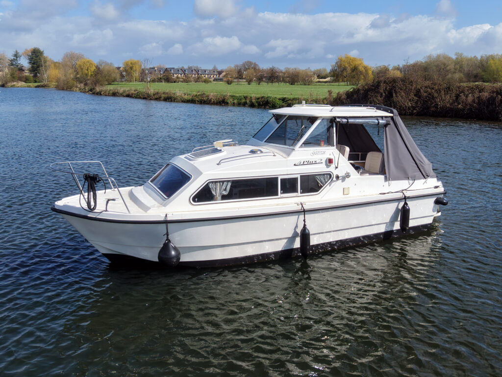 A white cabin cruiser boat floating on calm water with a canopy. The boat is moored near a green riverbank with trees. The sky is partly cloudy, providing a clear view of the surroundings.