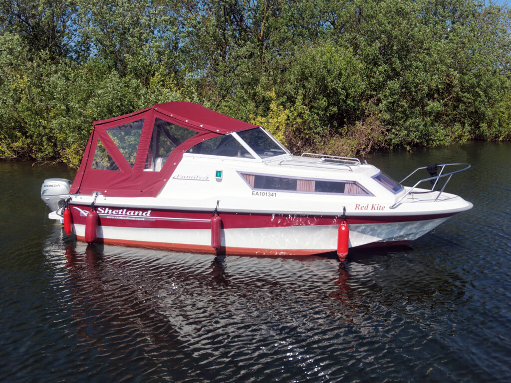 A white and red motorboat named 'Red Kite' anchored on still water surrounded by greenery. The boat features a red canopy and is equipped with a motor. Reflective water surface and trees line the bank in the background.