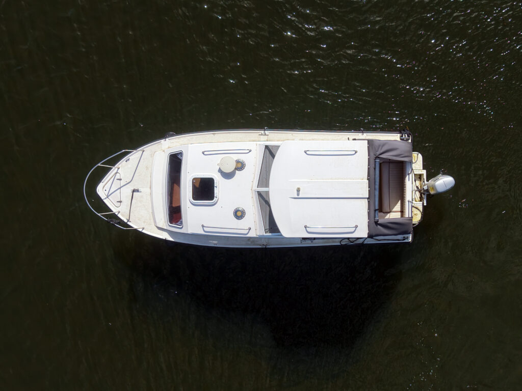 An aerial view of a white motorboat anchored on dark water. The boat features a spacious deck, a small seating area at the back, and a spotlight on the front. The boat appears stable, with the water reflecting the sunlight.