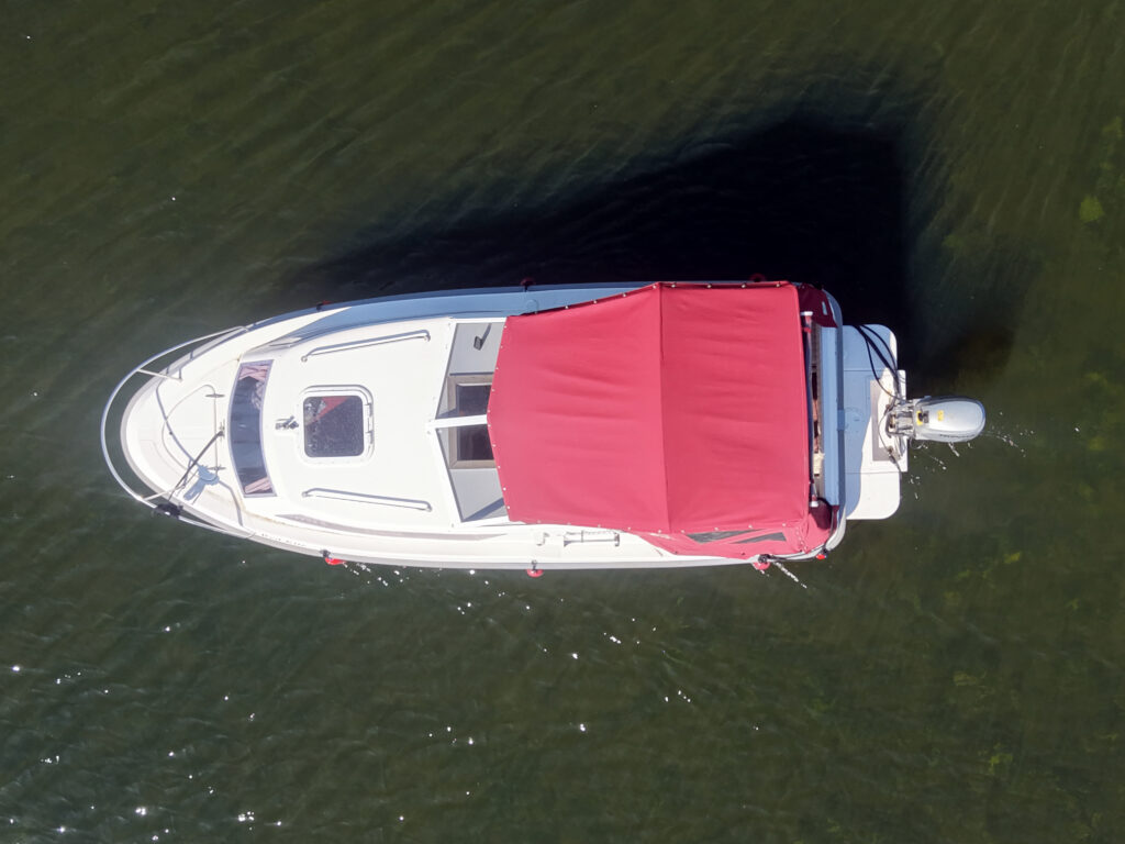 An aerial view of a white boat with a red canopy on the water. The boat's motor is visible at the rear, and the surface reflects the sunlight, creating a glimmering effect on the water.