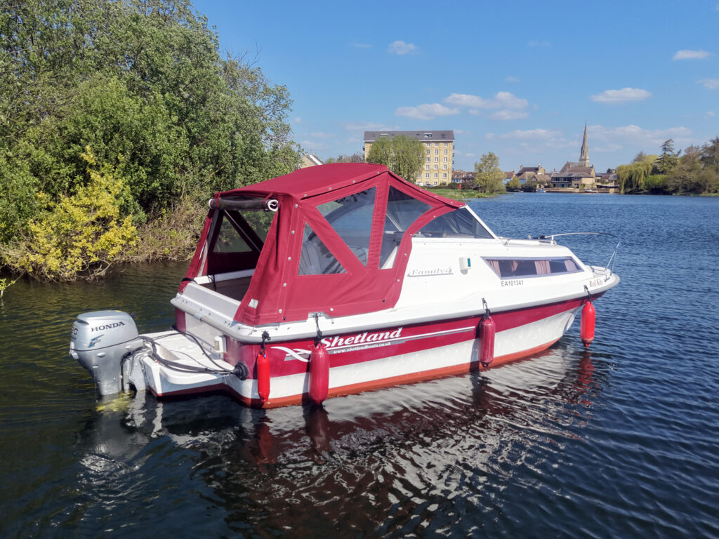 A white and red Shetland boat anchored on calm water. The boat features a red canopy and a Honda outboard motor. Surrounding it are green trees and a distant landscape with buildings and a church spire under a clear blue sky.