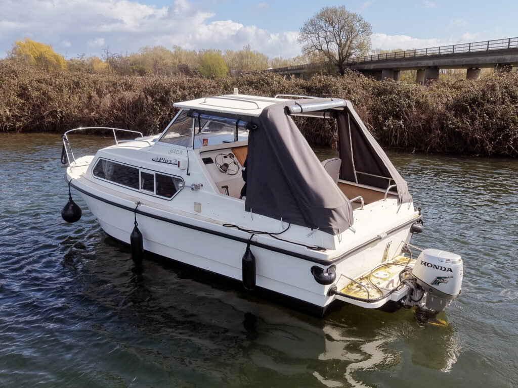 A white motorboat with a gray canopy is anchored on a calm river. The boat has a steering wheel and an interior seating area visible through the open cabin. In the background, a bridge and trees line the waterway under a partly cloudy sky.