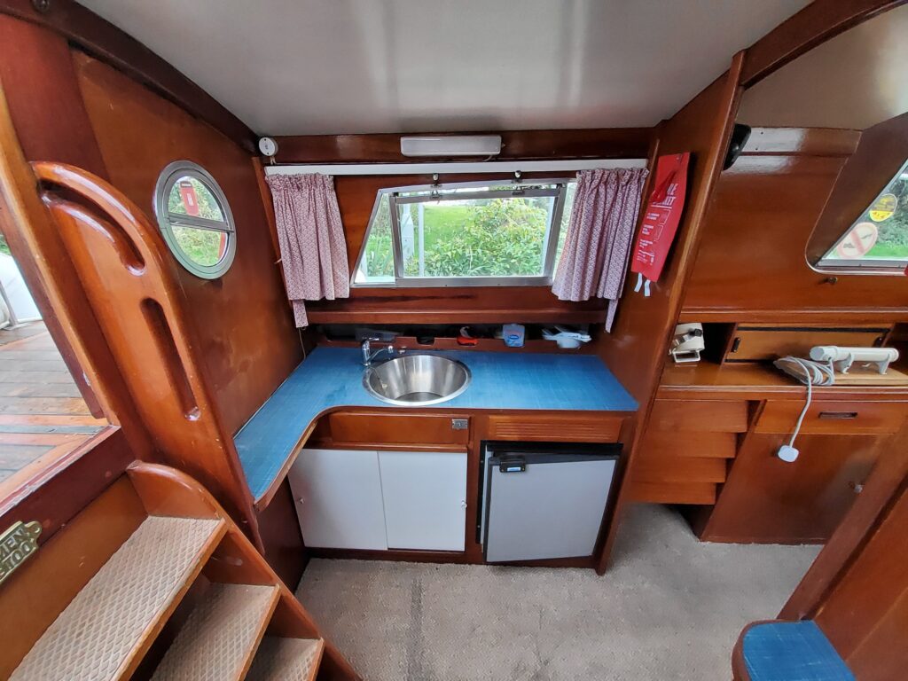 An interior view of a boat kitchen featuring wooden paneling, a stainless steel sink on a blue countertop, and a small refrigerator. The space includes curtains at the window and a staircase leading down, with storage visible on the right.