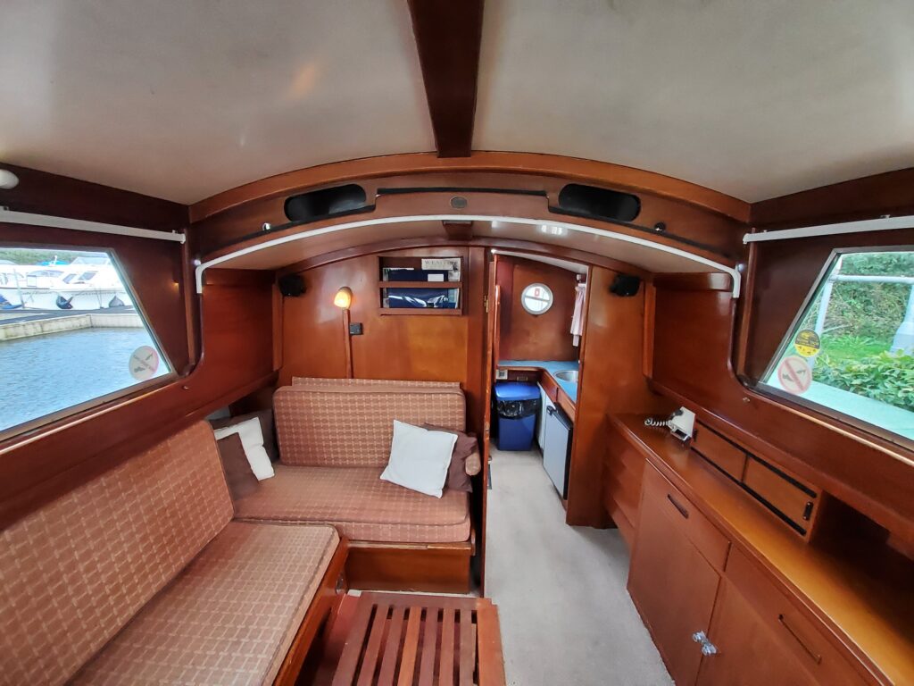 The interior of a boat features wooden paneling, with a seating area comprised of two cushioned couches and a wooden table. There is a small kitchenette area on the right, a clock on the wall, and large windows allowing natural light.