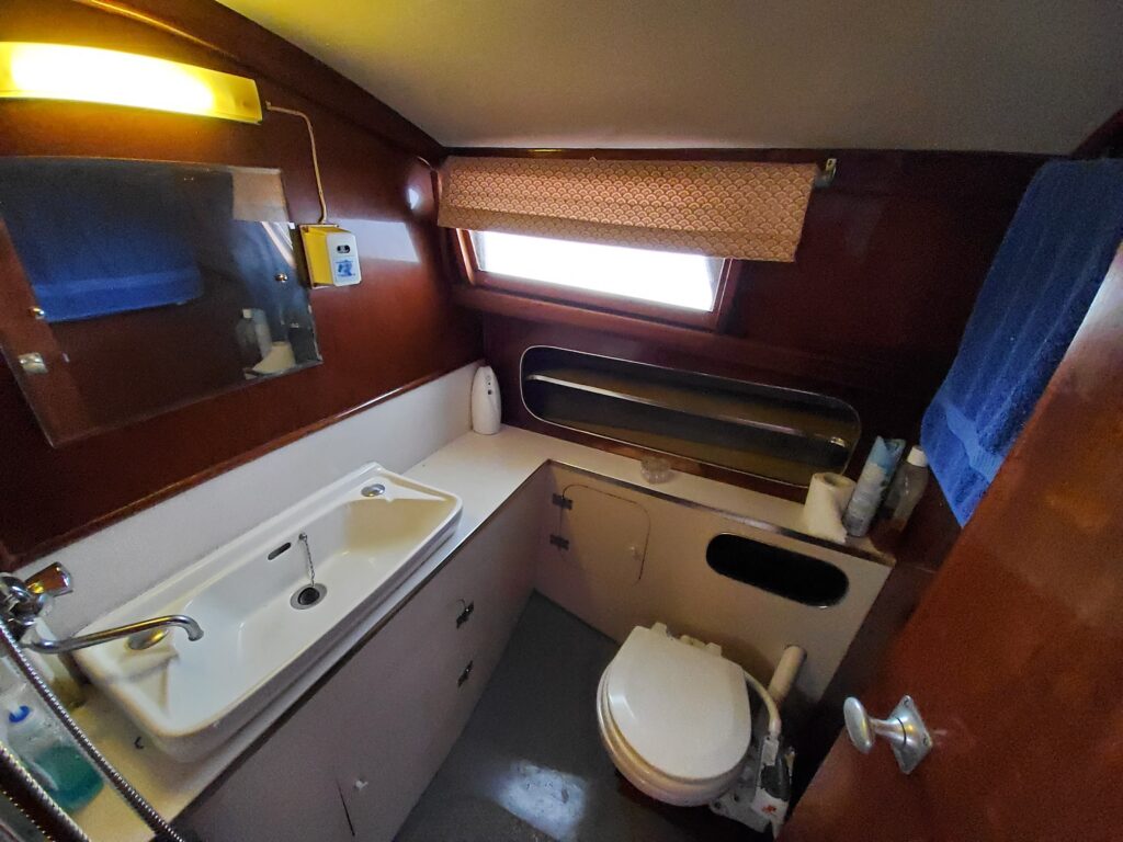 A small, enclosed bathroom on a boat featuring a compact sink with a faucet, a toilet, and a large window with curtains. The cabinetry is wooden, and toiletries are arranged on the countertop. Blue towels hang on the wall.