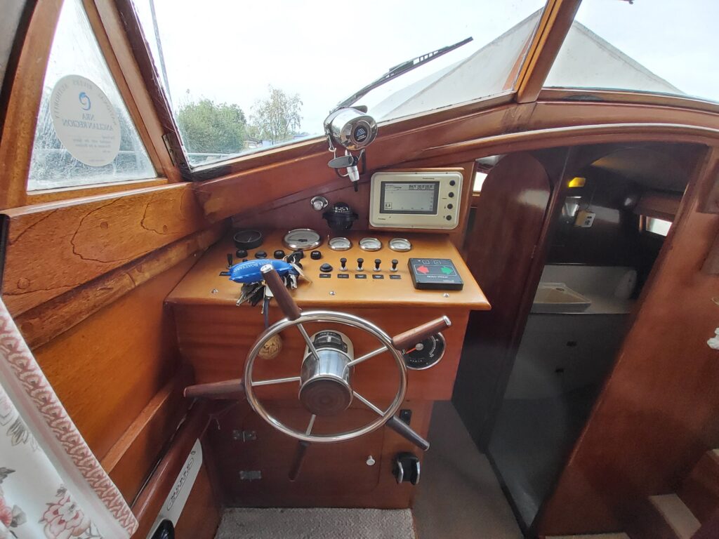 The interior of a classic L H Jones Cruiser featuring a wooden helm with a steering wheel, various control buttons, a gauge display, and a mounted compass. Natural light enters through windows, illuminating the space.
