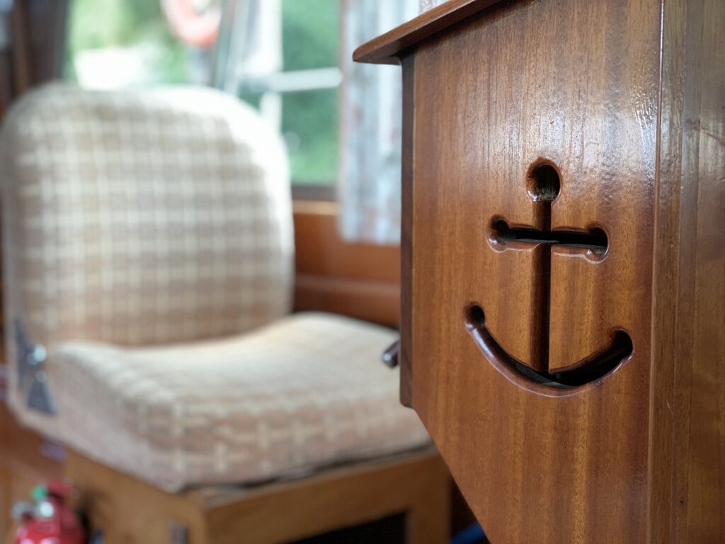 A close-up of a wooden cabinet with an engraved anchor design, beside a cushioned chair featuring a checkered pattern. Natural light enters through large windows in the background, illuminating the interior.