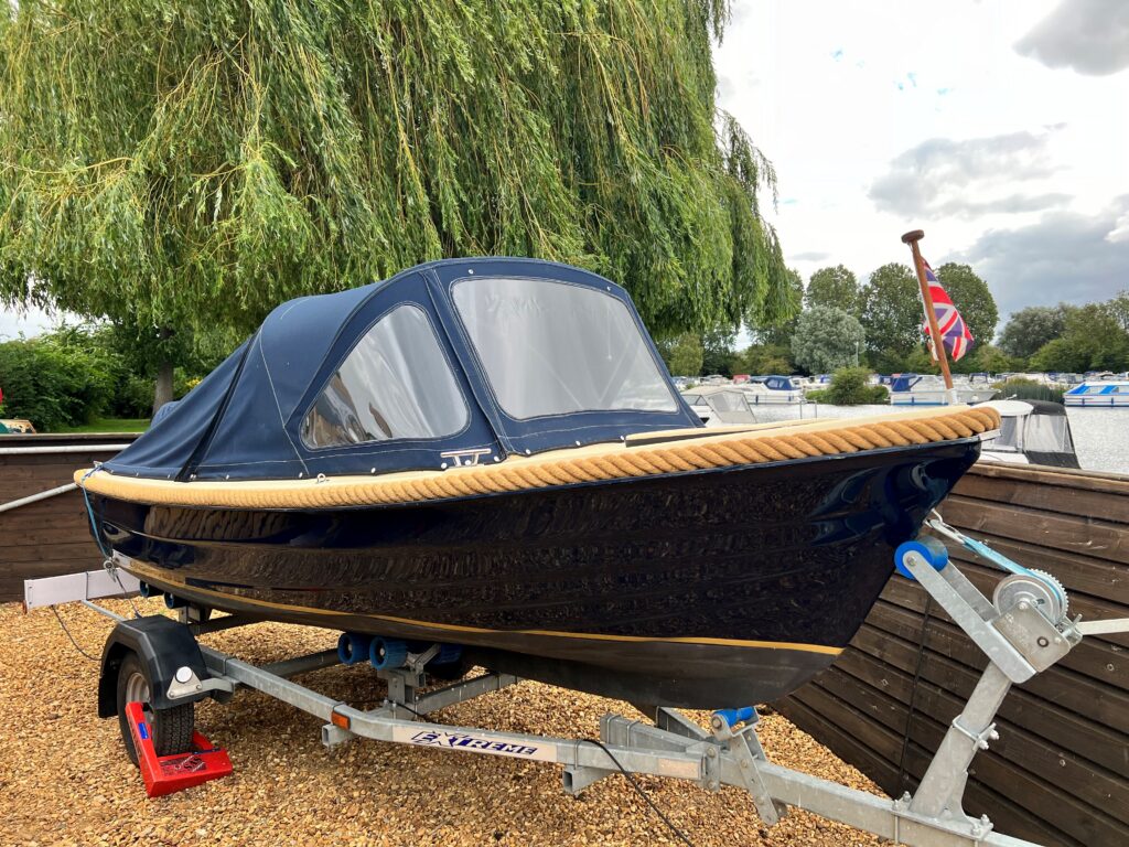 A dark blue boat with a canopy is mounted on a trailer and surrounded by gravel. The boat features a wooden trim along the edge and is positioned near a wooden fence with greenery and boats visible in the background.