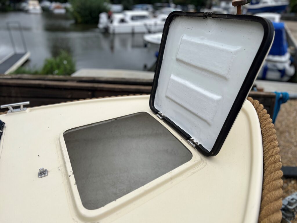 An open hatch on the top of a boat reveals a gray compartment, set against the backdrop of a marina with several boats moored in the water. The hatch has a white cover framed in black, and a rope coils along the edge of the boat.