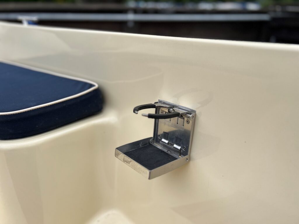 A close-up view of a stainless steel accessory holder mounted on a light-colored surface, featuring a small foldable shelf. A dark blue cushion is visible in the background.