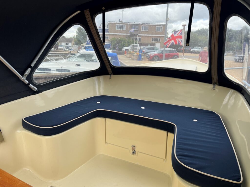 A seating area inside a boat, featuring a curved navy blue cushion with white trim. The background includes large windows with a view of a marina, a British flag visible outside, and boats parked in the distance.