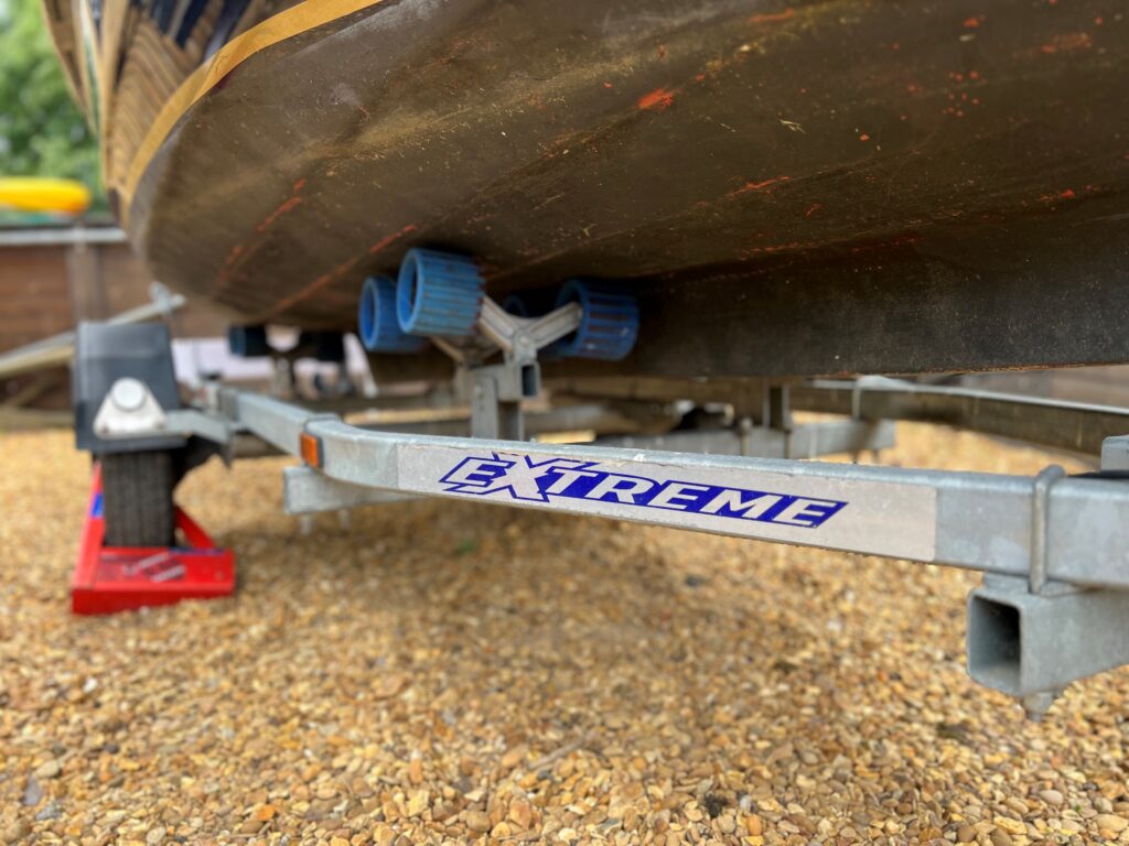 A close-up view of a boat trailer with the word "EXTREME" in blue letters. The trailer has rubber rollers and is positioned on a gravel surface, with part of a boat visible above, and a few objects in the background.