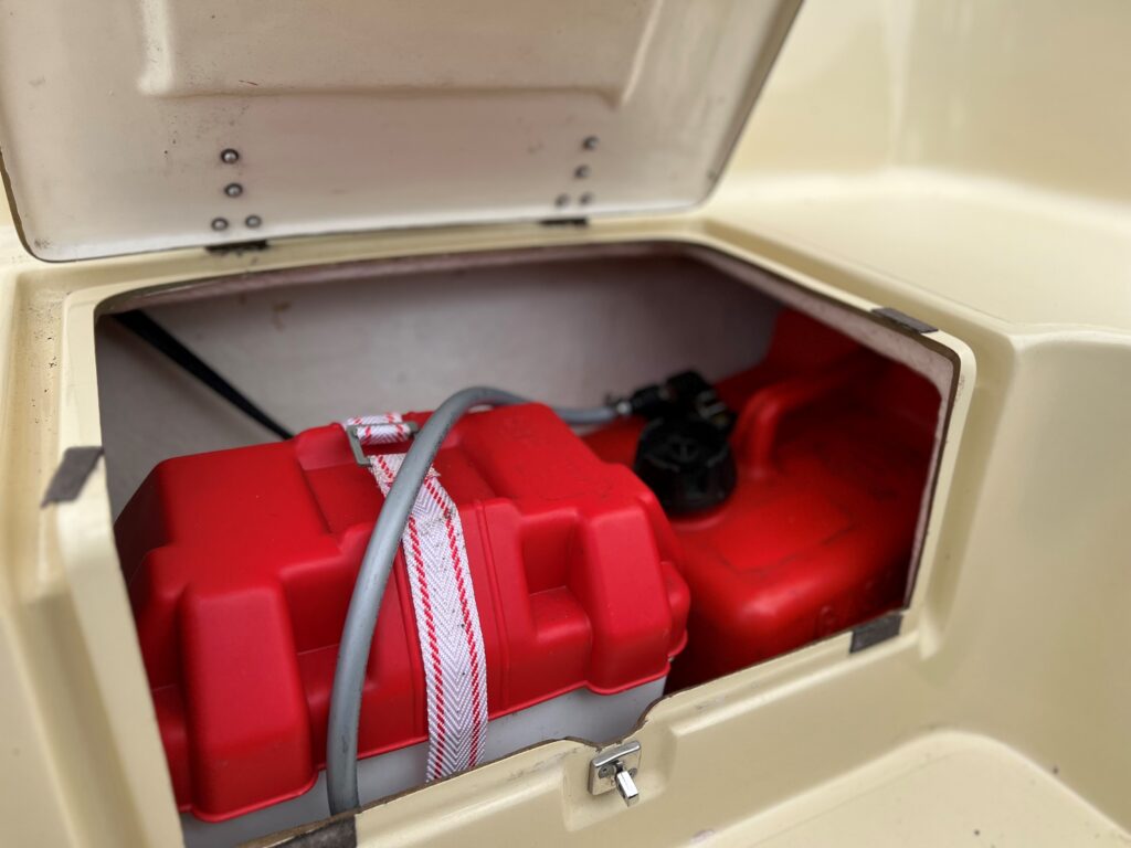 An open compartment reveals a red fuel tank secured with a strap and connected to a gray hose inside a beige surface. The compartment is rectangular, providing a view of the tank's lid and fittings.