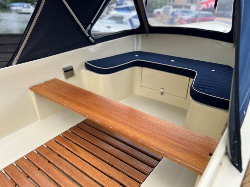 An interior view of a boat featuring a wooden plank bench and cushions in navy blue. The space includes a wooden floor and a compartment under the bench, with windows providing a view of the harbor and greenery outside.