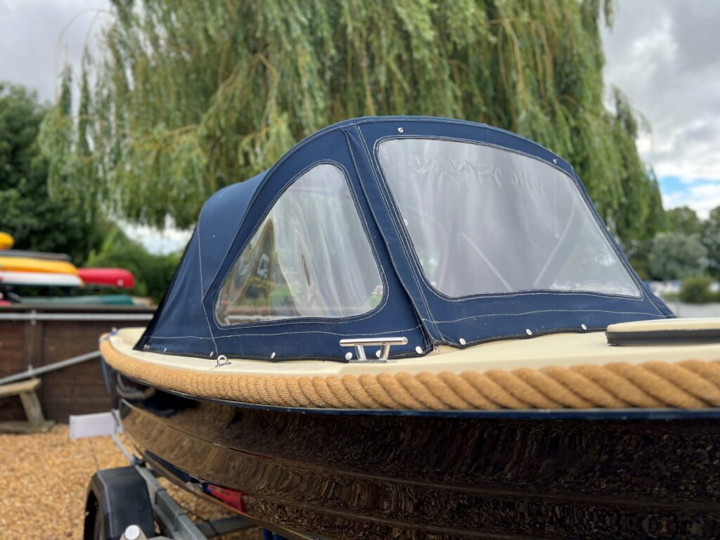 A close-up view of the bow of a boat featuring a navy blue canopy with transparent windows. The boat is moored on a pebbled surface, with greenery visible in the background and other boats partially seen behind it.