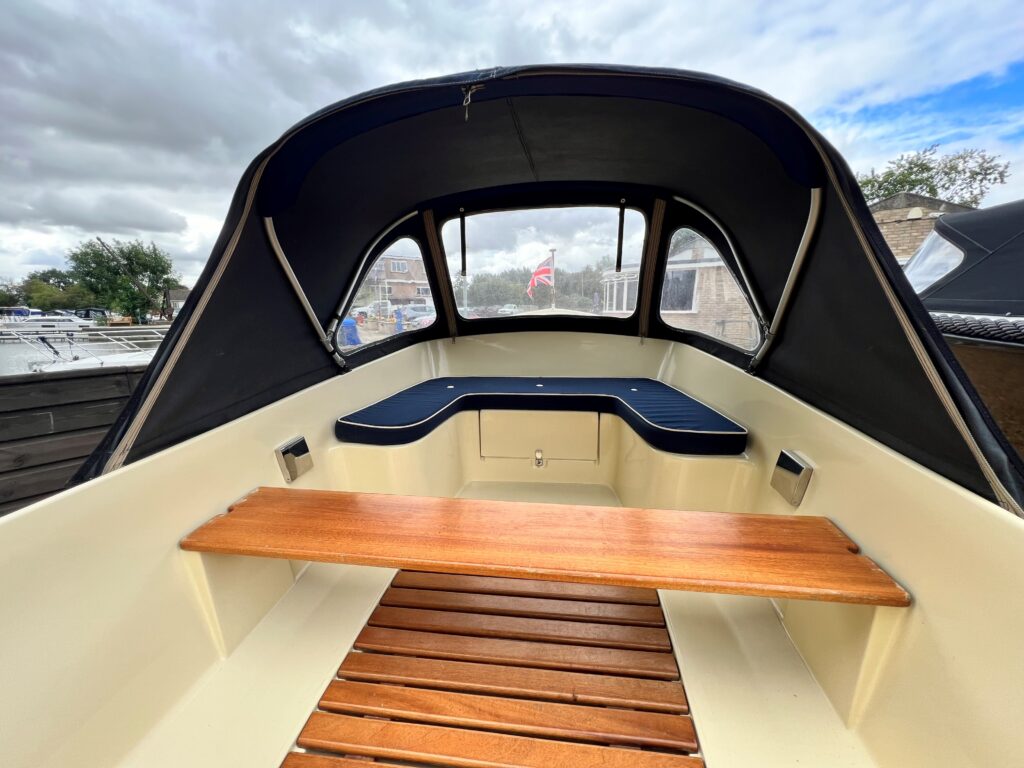 The interior of a boat featuring a wooden bench and a blue cushioned seating area. The cabin is enclosed with a dark canopy, and the surroundings include a view of a marina with boats and a cloudy sky.