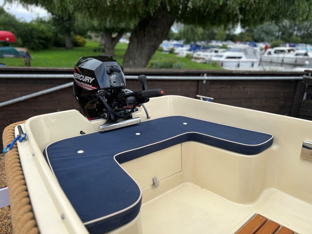 A close-up view of a boat's interior featuring a dark blue cushioned seating area surrounding an outboard Mercury motor. The background shows a marina with several parked boats and green trees.