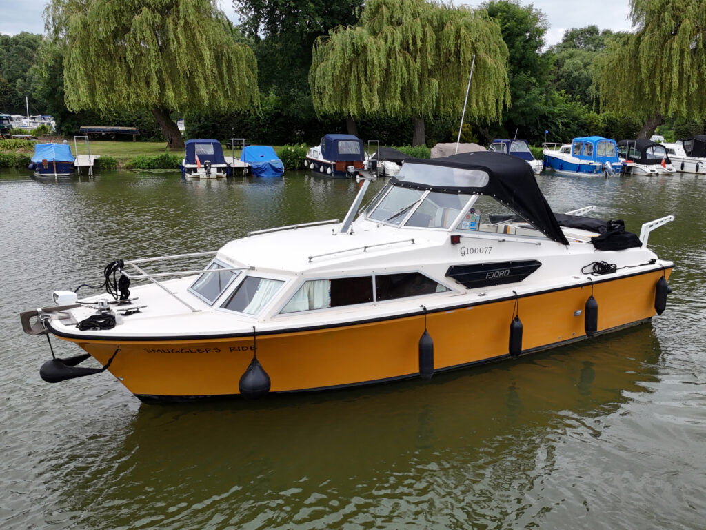 A Fjord Selcruiser 27 boat with a white and orange hull is moored in calm water. The boat features a black canopy and has several fishing rods secured on the front. Surrounding boats are visible in the background, along with lush greenery.