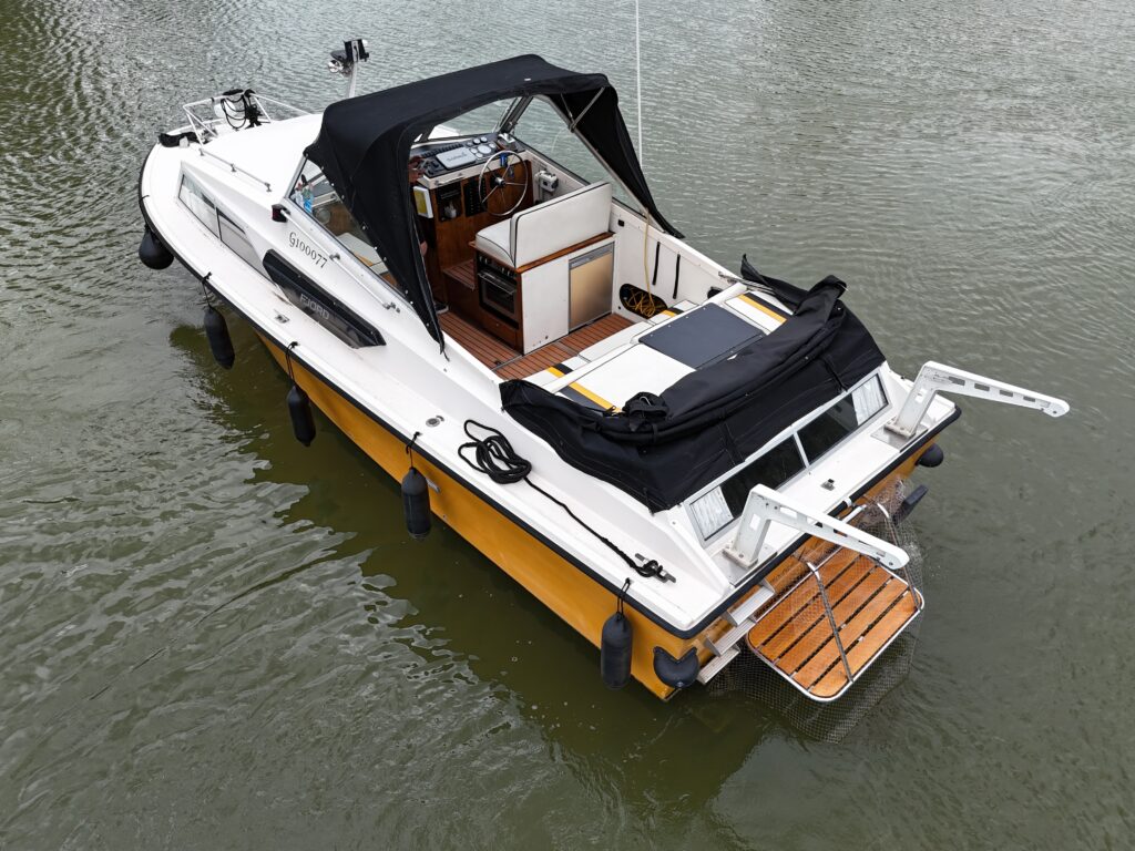An aerial view of a Fjord Selcruiser 27 boat on water. The boat has a white and orange hull with a black canopy covering the cockpit area. The rear features a wooden platform, and the interior layout includes seating and navigational equipment.