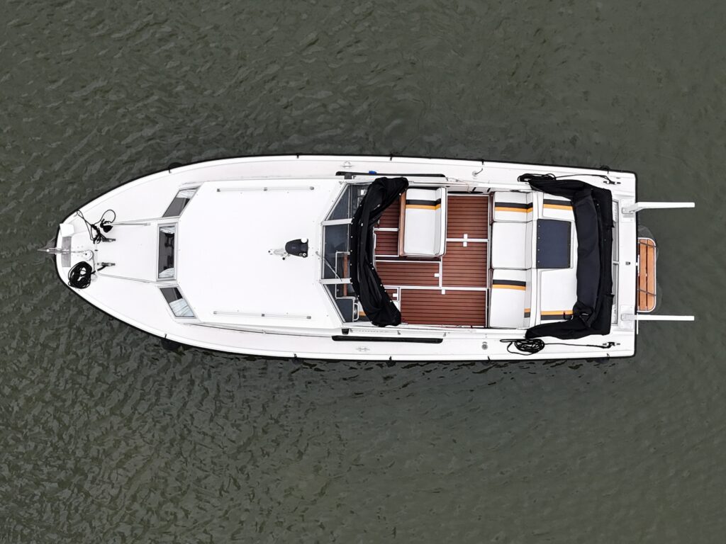 An overhead view of a white boat with a sleek design featuring a deck made of wood slabs. The boat has black upholstery around the seating area and a small table in the center, resting on calm water.