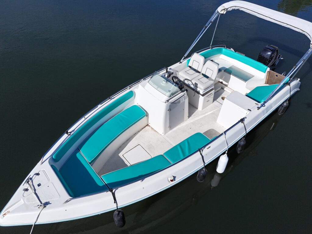 A white motorboat with turquoise cushions is anchored on calm water. The boat features a spacious seating area with two front chairs and a console in the center, surrounded by cushioned benches along the sides.