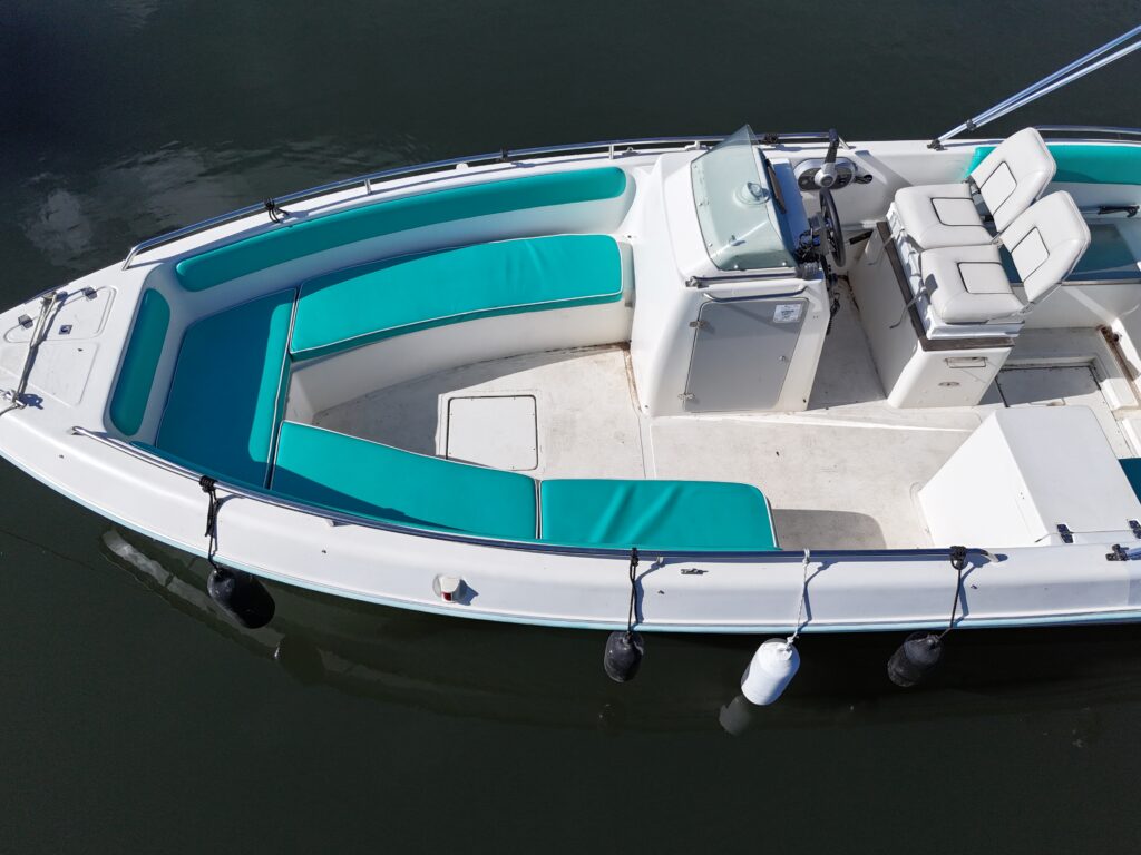 An aerial view of a white boat featuring teal cushioned seating along its sides. The boat has a center console with a steering wheel and two adjacent seats, and it is docked in calm water.