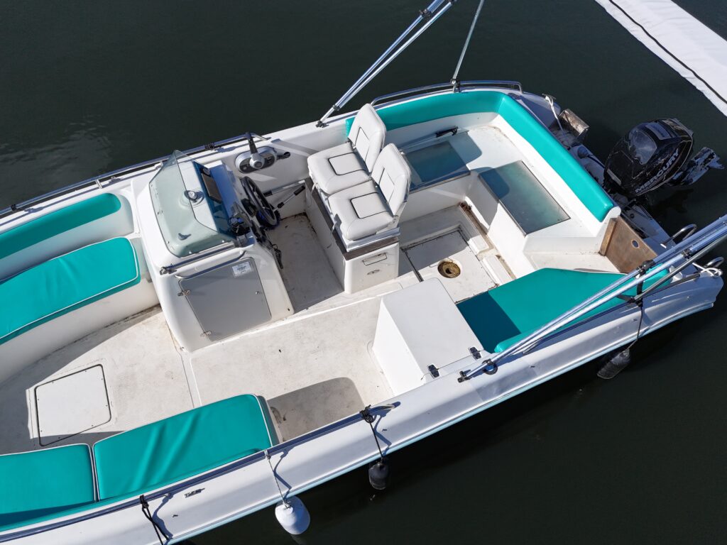 A top-down view of a boat featuring teal cushions on white seating. The interior includes a steering wheel, two captain's chairs, and a central console, with an outboard motor visible at the back. The boat is moored with a dock visible in the background.