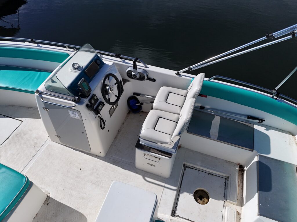 An overhead view of a boat's interior featuring a center console with a steering wheel, throttle controls, and a seat beside it. The bright, teal and white seating cushions contrast with the boat's white flooring, and the calm water is visible in the background.