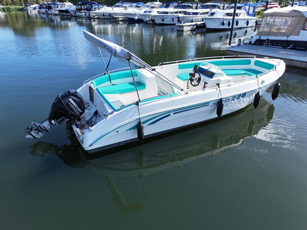 A white motorboat with teal accents is moored in calm water at a marina. The boat features a steering wheel and an outboard motor, surrounded by other boats docked nearby.