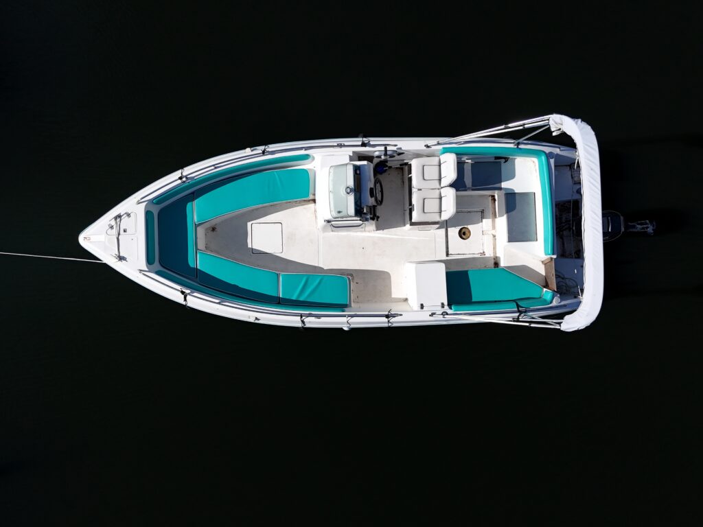 An aerial view of a white boat with teal accents anchored on dark water. The boat features a spacious deck with cushioned seating and a console at the front, creating a modern design ideal for leisure activities.
