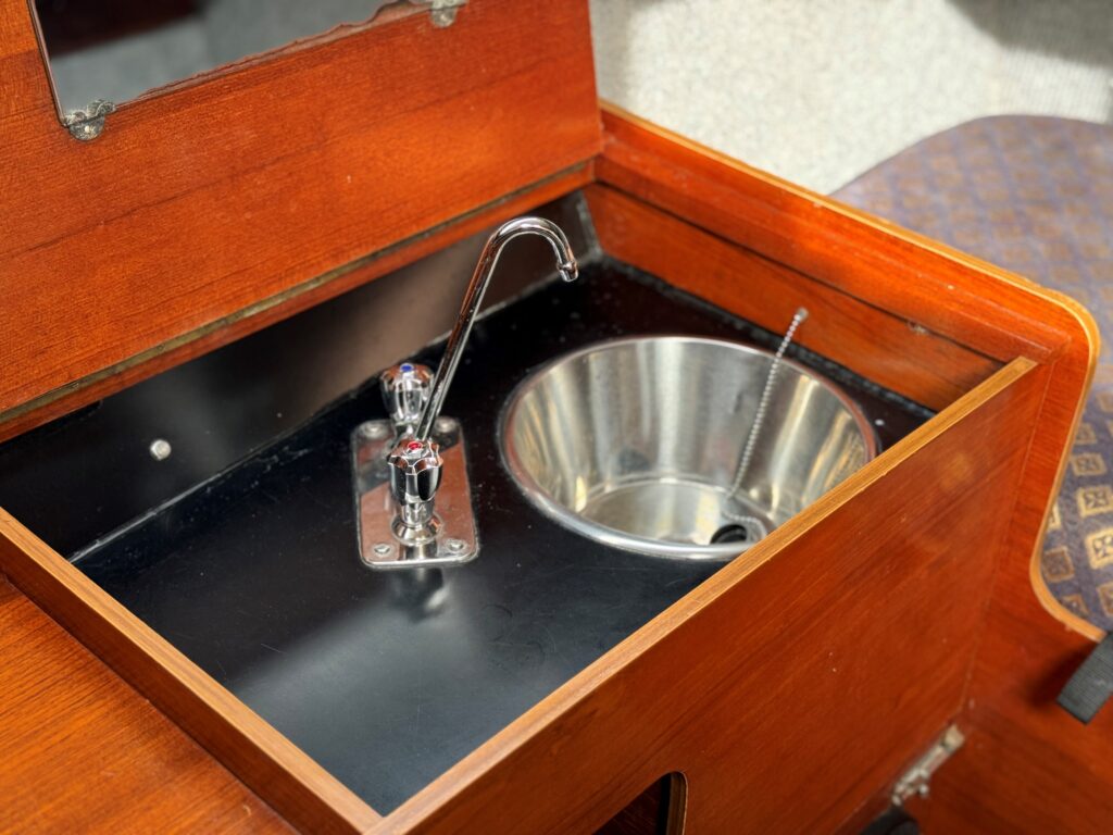 A wooden countertop features a stainless steel sink and a faucet. The sink is circular with a polished surface, and the faucet is installed on a metal plate next to it. The background includes a patterned cushion and a wall with a mirror.