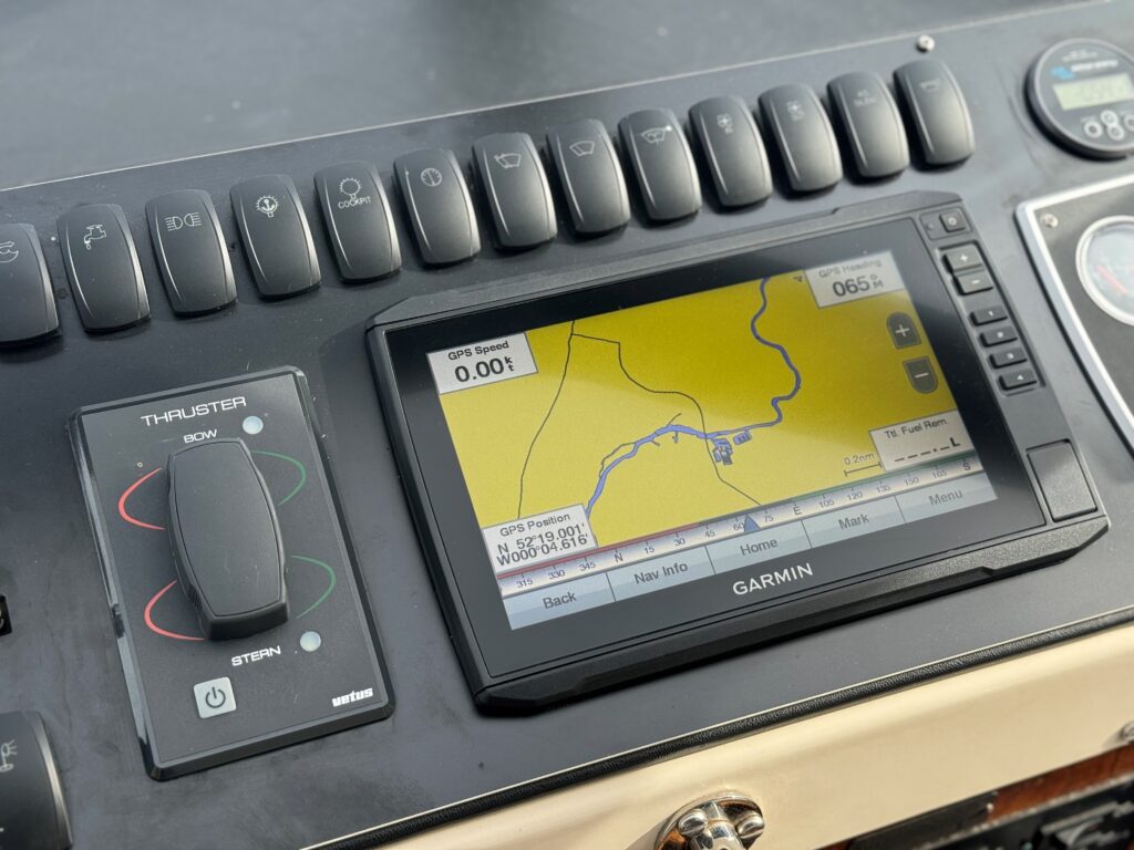 A close-up view of a boat's control panel featuring a Garmin GPS device displaying a map with a yellow background. Nearby are navigational buttons and a thruster control lever, all set against a wooden surface.