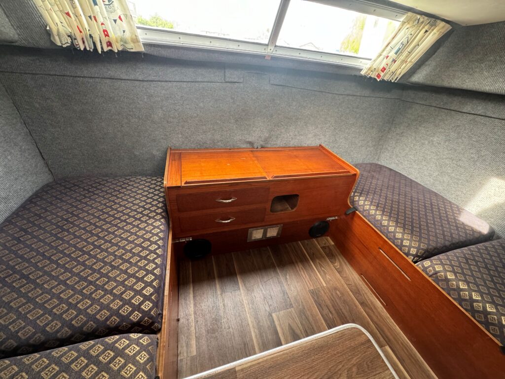 An interior view of a boat featuring two upholstered seating areas with patterned cushions, a wooden table with two drawers in the center, and a window overhead bringing in natural light. The floor is made of wooden planks.