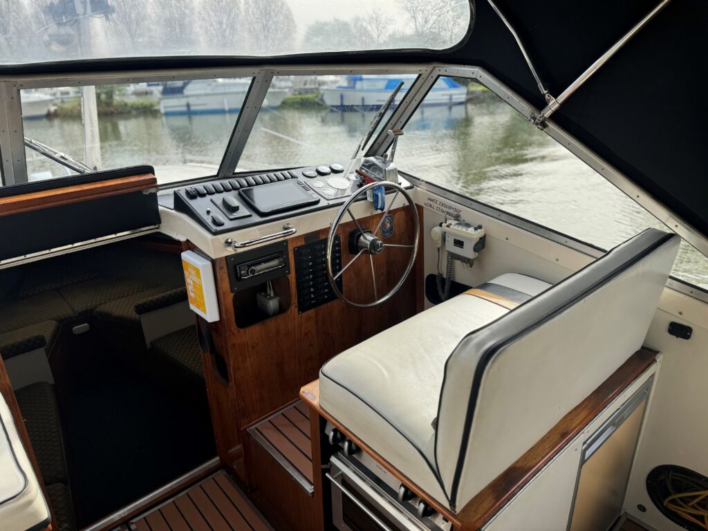 The interior of a boat featuring a wooden steering wheel, control panel, and a beige cushioned captain's seat. The dashboard includes various instruments and buttons. Visible through the windows are boats and trees by the water.