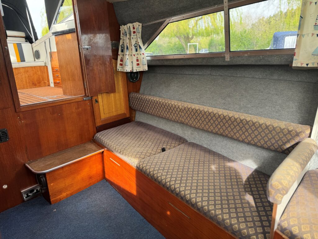 The interior of a boat features a brown upholstered bench seat with a patterned fabric. A wooden panel and two small windows provide natural light, while a door leads to the outside. The floor is covered in blue carpet.