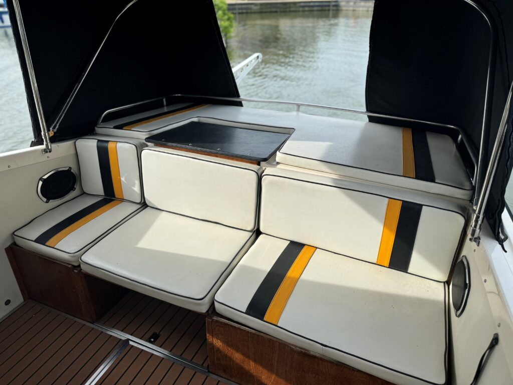 The interior seating area of a Fjord Selcruiser 27 boat features cushioned benches with white upholstery accented by yellow and black stripes. A small table is positioned at the center, with a view of the water visible through the open rear.