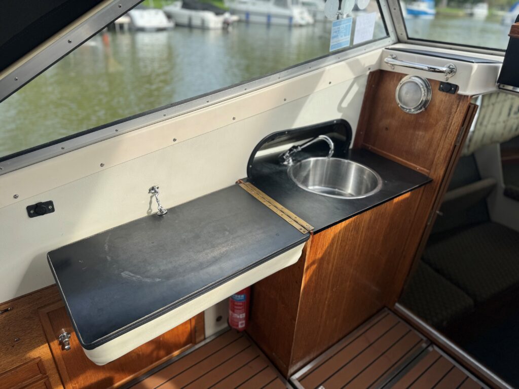 The interior of a boat showing a wooden kitchenette with a stainless steel sink and a black countertop. The counter has a small faucet and a wooden section, while the background features a view outside the boat with moored vessels and green surroundings.