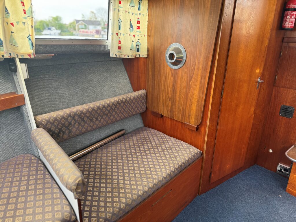 An interior seating area of a boat featuring a cushioned bench with a patterned fabric along the wall. There's wood paneling on the walls and a round window on the right. The area has curtains decorated with lighthouse designs, and the floor is covered with blue carpet.