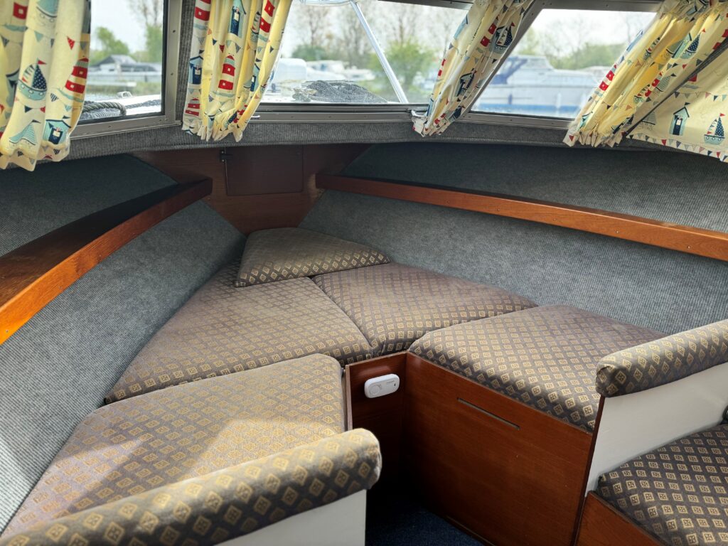 The interior of a Fjord Selcruiser 27 boat features a cozy seating area with brown and beige patterned cushions. The walls are lined with gray fabric, and colorful curtains adorned with nautical motifs frame the large windows, allowing natural light to enter.