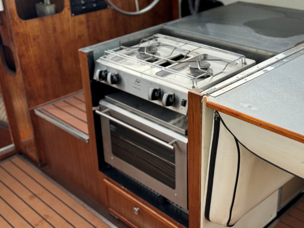 A stainless steel stove and oven unit installed in the kitchen of a boat. The unit features two gas burners on top and an oven below, set against wooden cabinetry and a countertop with a silver finish.