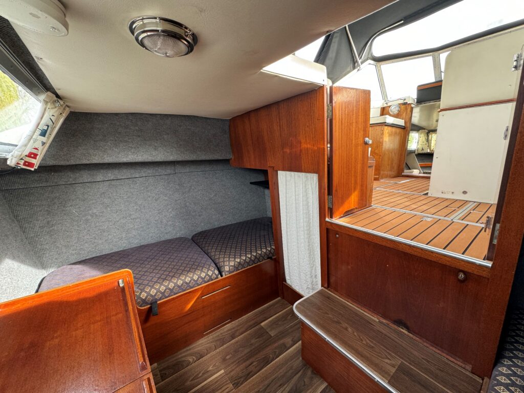 The interior of a Fjord Selcruiser 27 features a wooden bench with navy-patterned upholstery, adjacent to a cabin wall with gray fabric. A staircase leads to the main area, and the floor is covered in wooden planks.