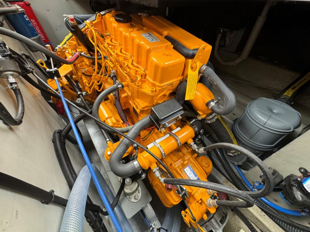 An orange marine engine is visible with several black and gray hoses connected to it. The engine is mounted on a boat's interior platform, surrounded by various components, including a gray cylindrical tank and electrical wiring in the background.