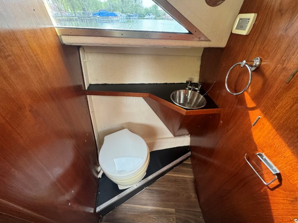 A compact boat bathroom featuring a white toilet, a small sink on a wooden shelf, and a mirror. The interior walls are wooden, and there is a window allowing natural light. The floor is a dark laminate, and a door leads outside.