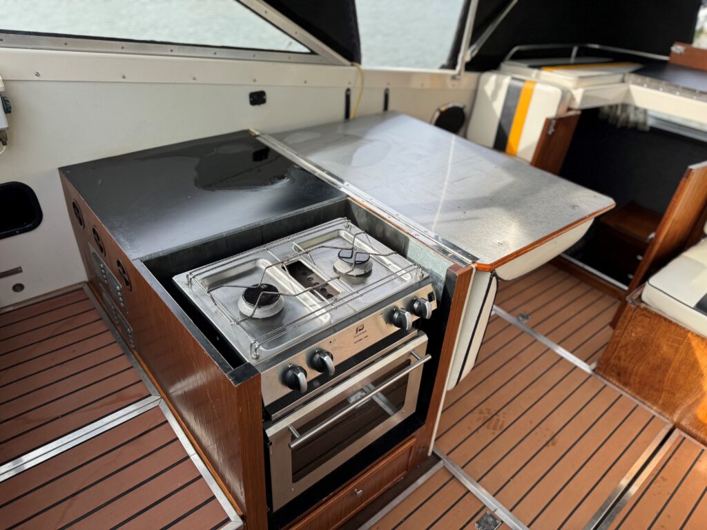 A compact kitchen area in a boat featuring a two-burner gas stove and an oven beneath, set into a wooden cabinet. A stainless steel countertop extends alongside, with a warm wood floor underneath.