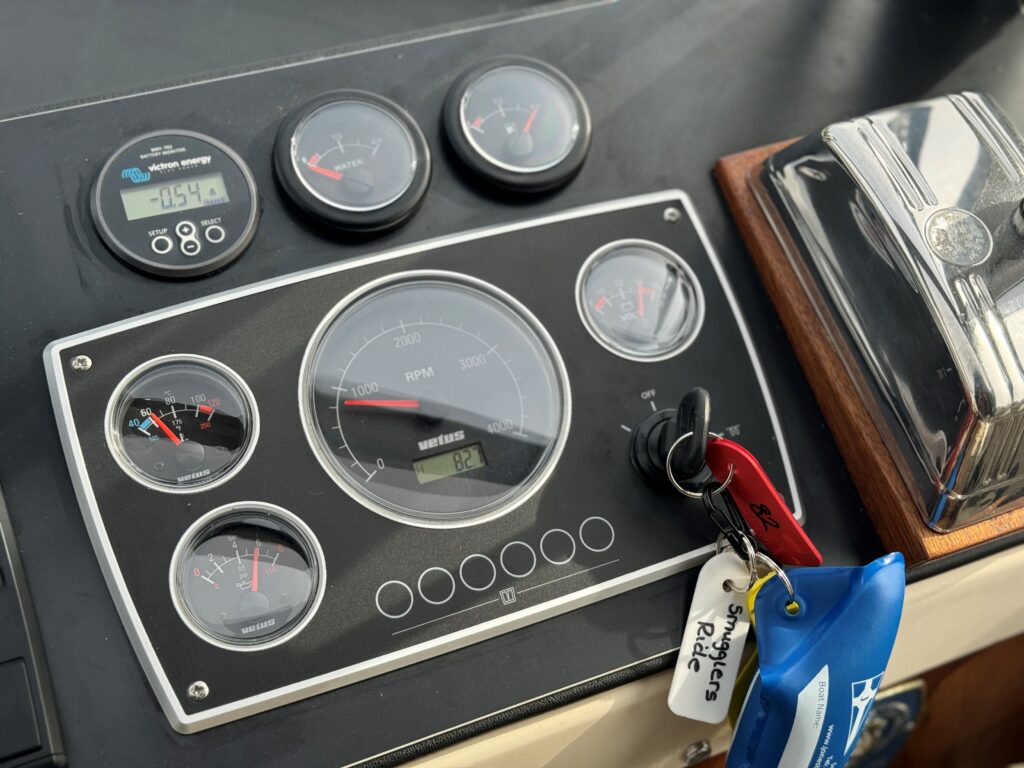 A close-up view of a boat's control panel featuring five circular gauges displaying various readings, a digital speedometer, and a key ignition with multiple keys attached. The panel has a sleek black finish and is positioned on a wooden surface.