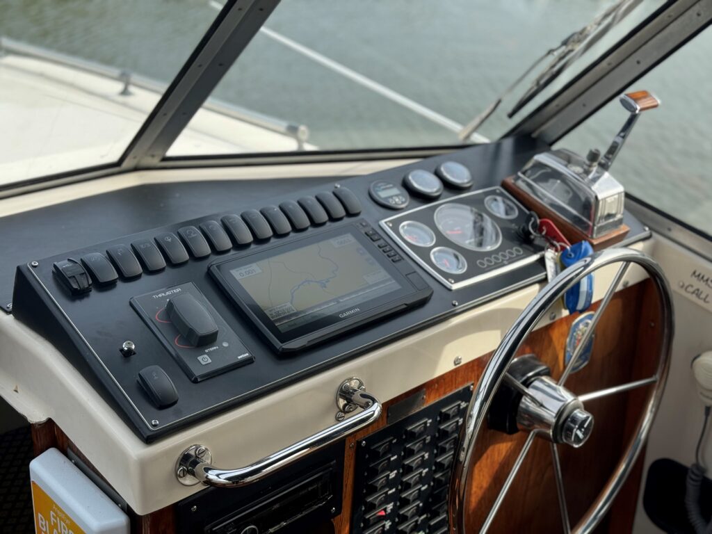 The cabin of a Fjord Selcruiser 27 boat featuring a navigation console with a touchscreen display, multiple dials, and control switches. A steering wheel is visible on the right side, while the background shows a view of the water through large windows.