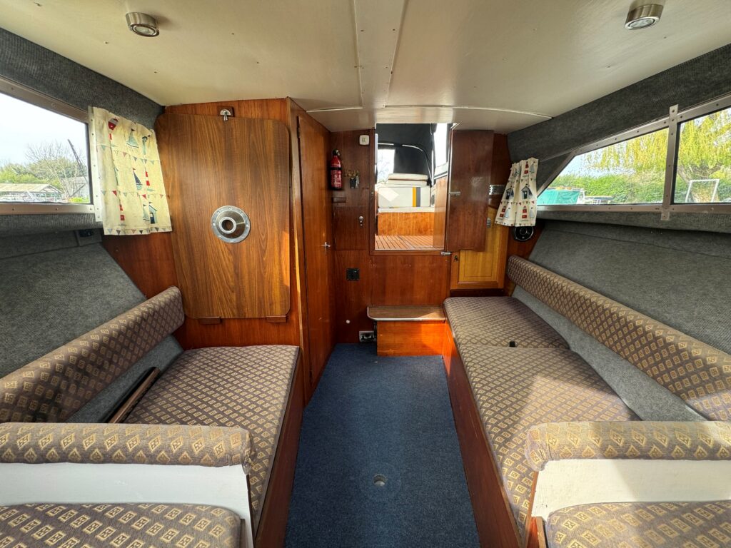 The interior of a Fjord Selcruiser 27 boat featuring wooden paneling, two upholstered benches with patterned fabric facing each other, and a small table in between. Large windows allow natural light, and the floor is covered with blue carpeting.
