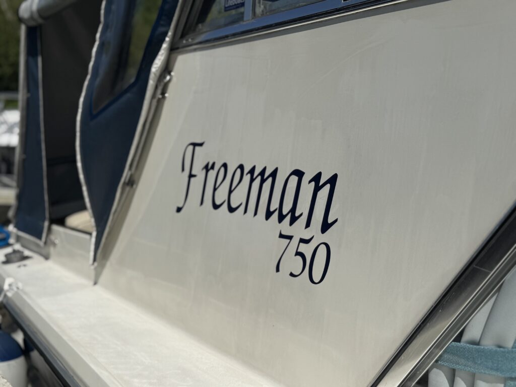 A close-up view of the side of a boat featuring the name "Freeman" in elegant script followed by the number "750." The boat's surface is white, with some background elements suggesting a marina setting.
