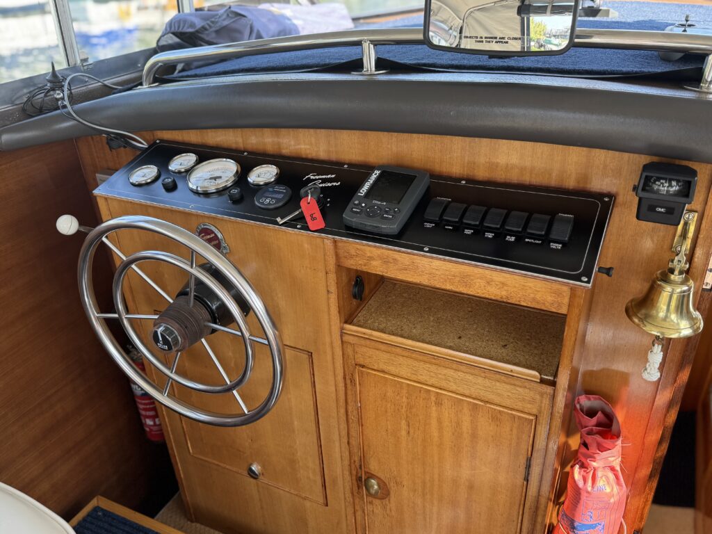 The interior of a boat showing a wooden control panel with various gauges, a steering wheel on the left, and a black electronic device with buttons. A bell hangs next to a small storage compartment with orange safety equipment below.