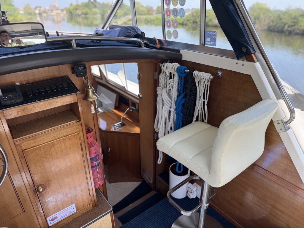 The interior view of a boat with a wooden dashboard featuring a control panel, a bell, and storage. A white swivel chair is positioned beside a bundle of ropes, and a window reveals greenery outside. The floor has a striped rug.
